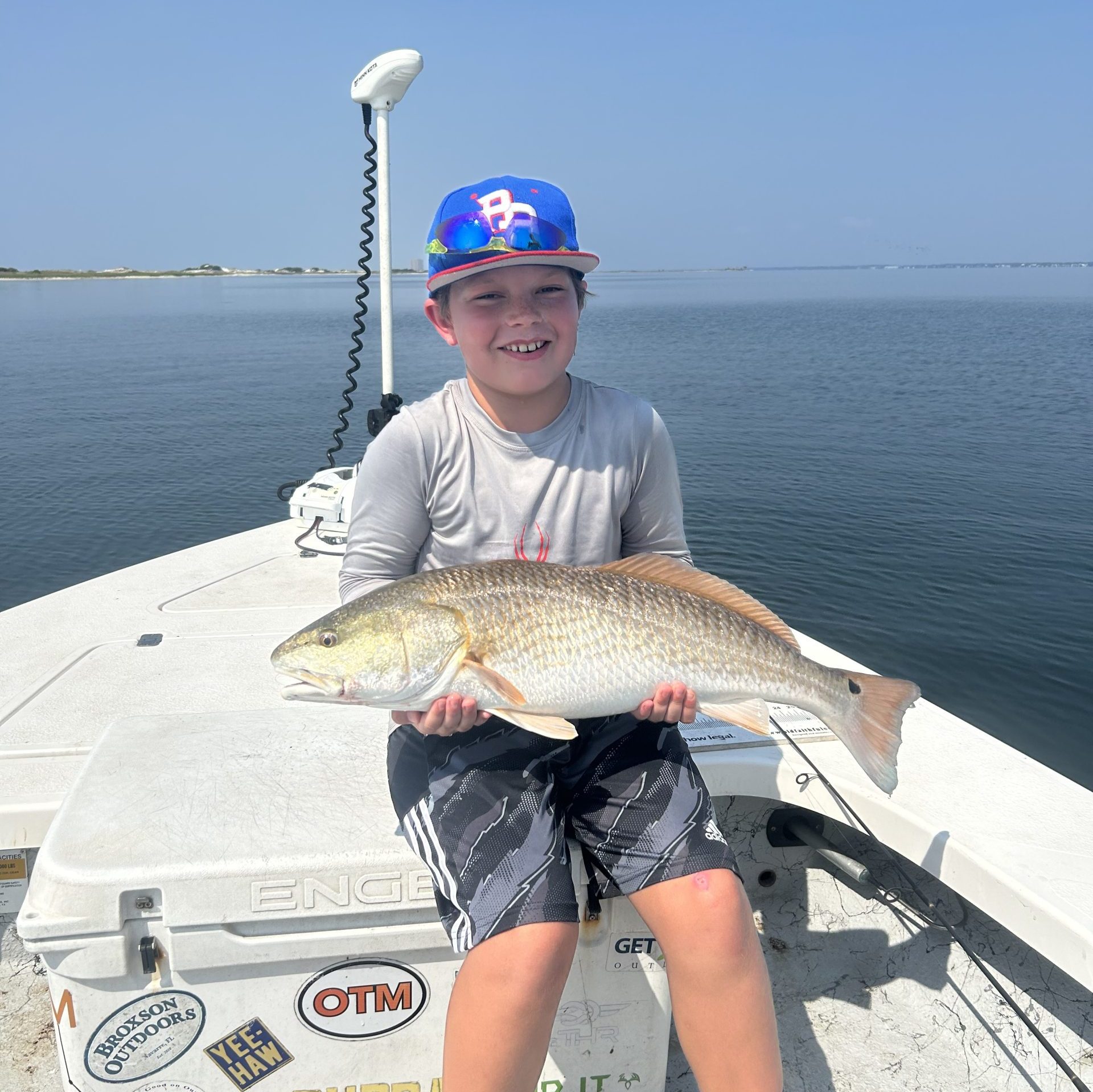 A young boy sitting on a professional fishing cooler holding a healthy Redfish caught in Pensacola Bay.