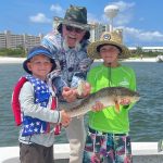 A grandfather and two grandsons holding a Redfish at the Navarre Beach Boat Ramp with the Navarre Beach Water Tower in the background during a family fishing charter.